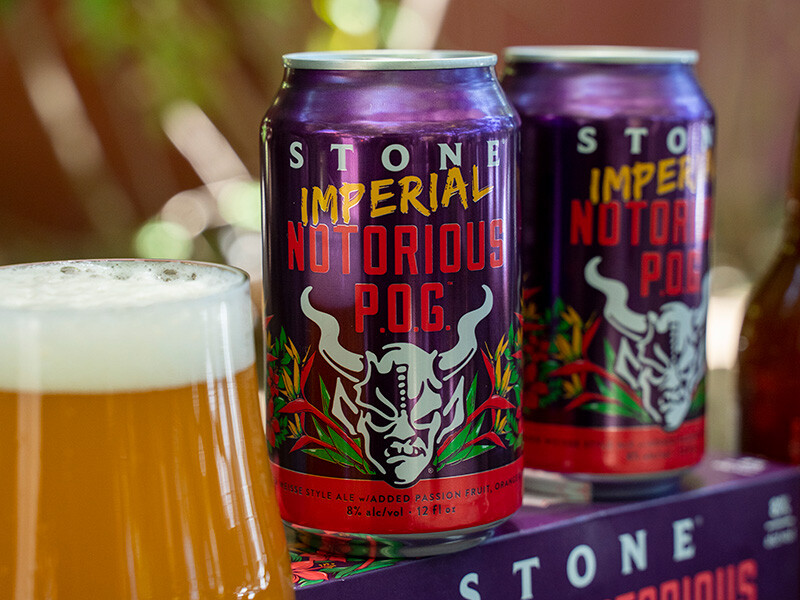 cans of Stone Imperial Notorious P.O.G. behind a glass of the juicy, hazy beer inspired by the hawaiian fruit drink