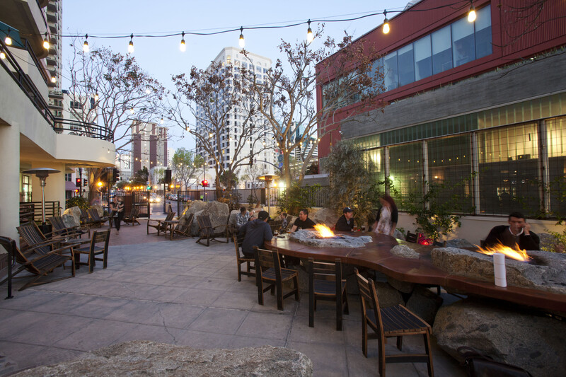 Guests gather around a long wooden table with built-in boulder fire pits on the Stone Brewing Tap Room - Kettner string-lit outdoor patio at dusk, with downtown high-rises in the background