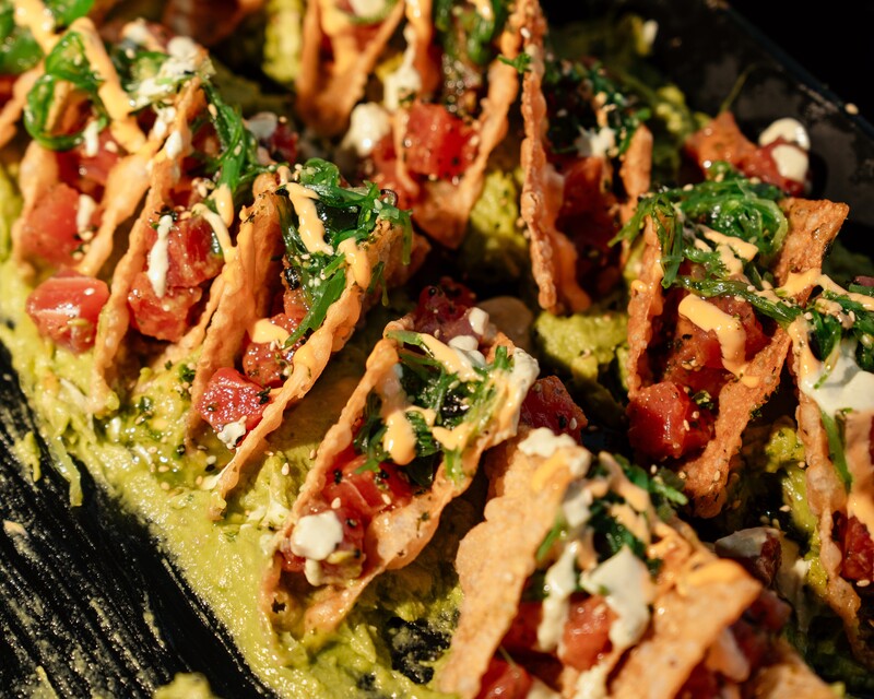 Rows of mini crispy wonton tacos filled with diced ahi tuna, seaweed salad, sesame seeds and a spicy mayo drizzle, set on a bed of guacamole