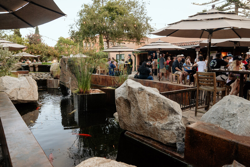 Koi swim in a rock-lined pond with a small waterfall beside a busy, umbrella-shaded Stone Brewing patio at dusk