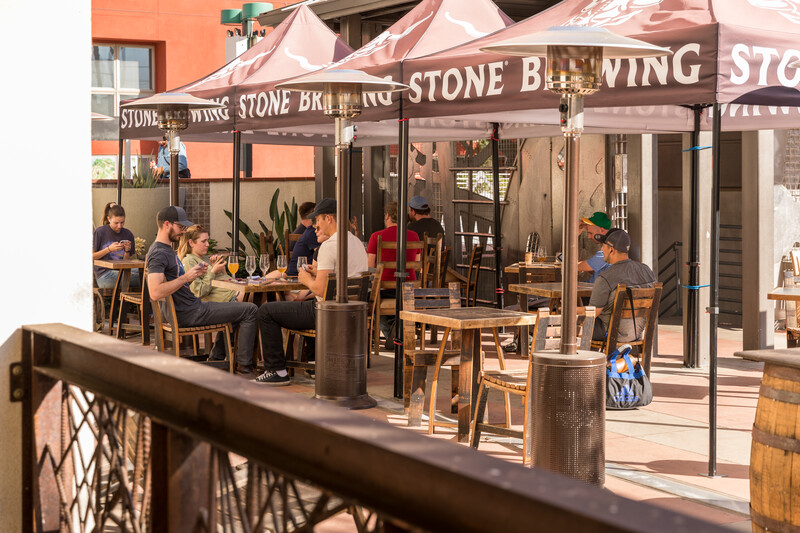 Friends play cards over beers at wooden tables under Stone Brewing canopies on the sunny Stone Pasadena patio