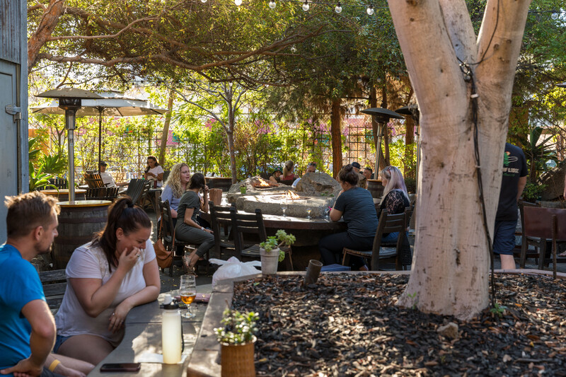 Guests relax at tables and around boulder fire pits on the tree-shaded Stone Oceanside patio on a sunny afternoon