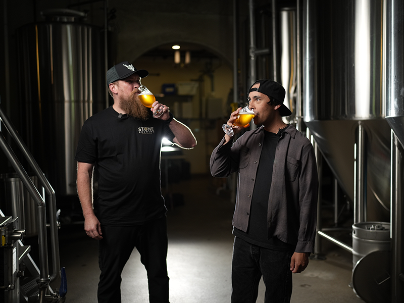 Vic and Kris Ketcham taste Stone Pierce the Veil IPA inside a brewery surrounded by stainless steel brewing tanks.