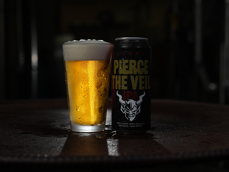 A glass of golden beer sits beside a can of Stone Pierce the Veil IPA on a table.