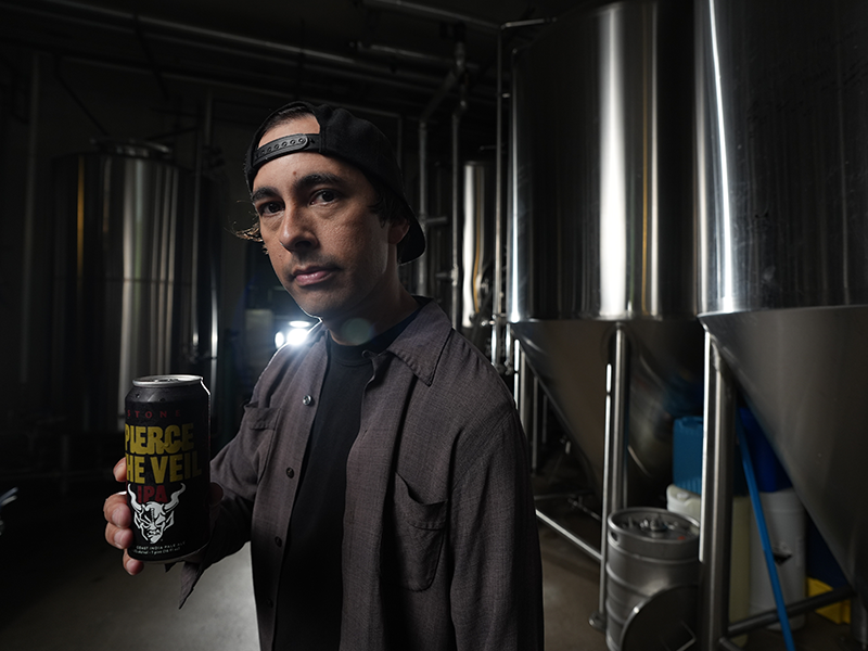 Vic holds a can of Stone Pierce the Veil IPA inside a brewery with stainless steel tanks in the background.