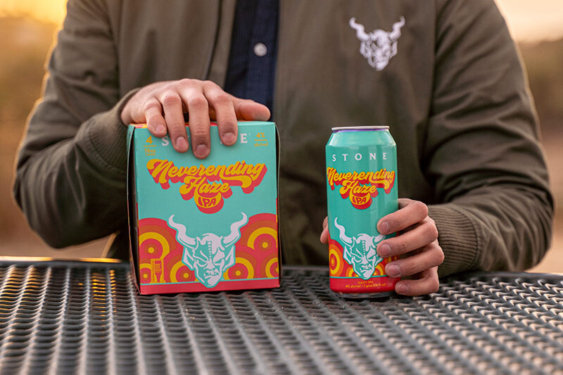 Stone Neverending Haze IPA can and four-pack on a table in front of a person